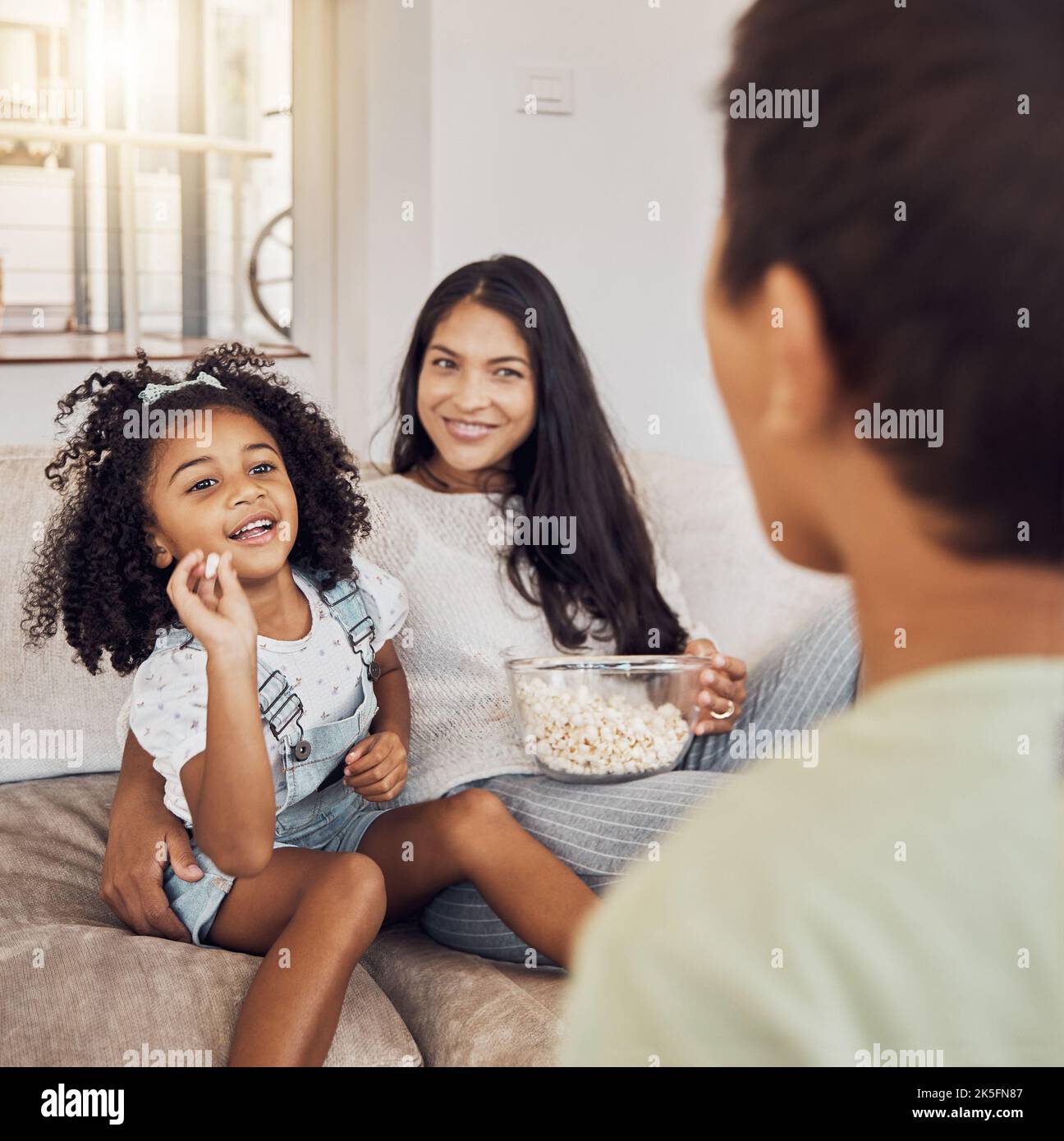 Mother, family and girl throw popcorn on sofa in home living room. Love, relax and child or kid with parents feeding food to person, spending time Stock Photo