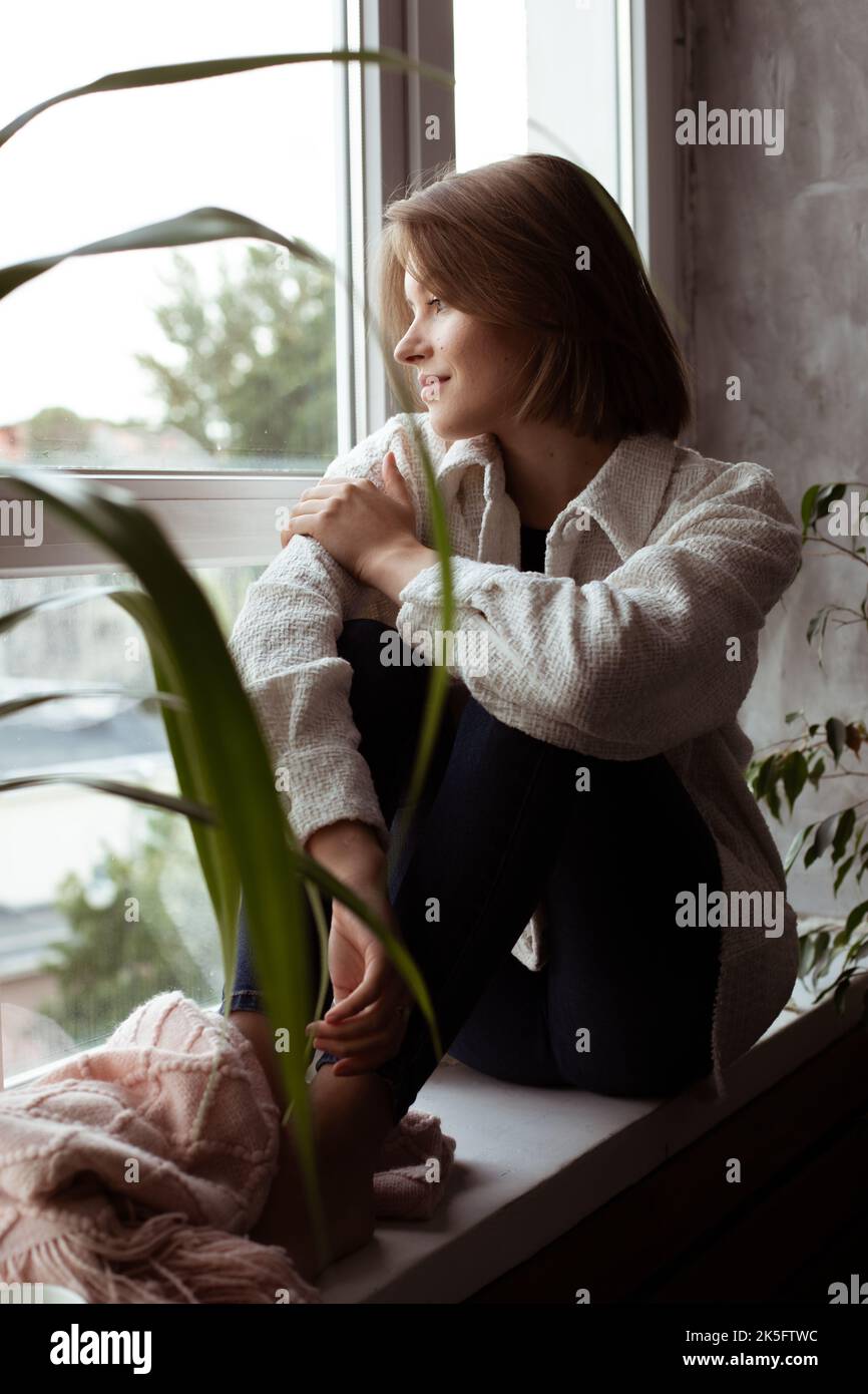 Vertical smiling calm pensive young brunette woman look out window, sit on windowsill in warm shirt. Dream of own house Stock Photo