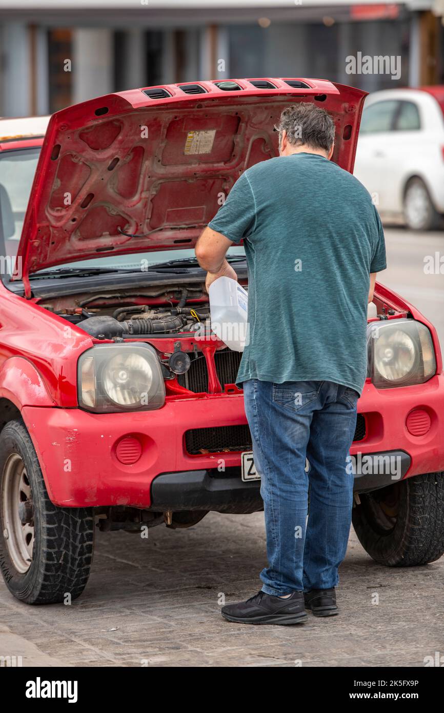 man standing next to car with open bonnet fixing the engine adding water to a car radiator to prevent overheating, car engine overheating add water. Stock Photo