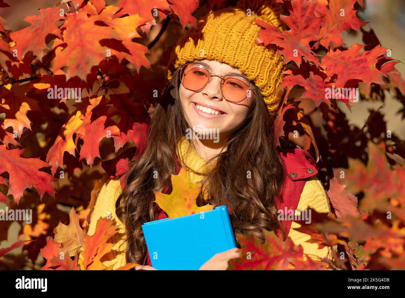 Autumn kid girl face in autumn fall leaves outdoor. glad kid hold book