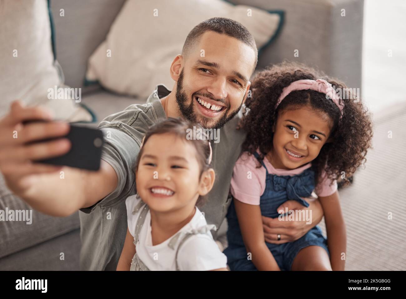 Father taking a selfie on his phone with his children hugging and bonding in their family home. Multicultural father and daughters smiling, happy and Stock Photo