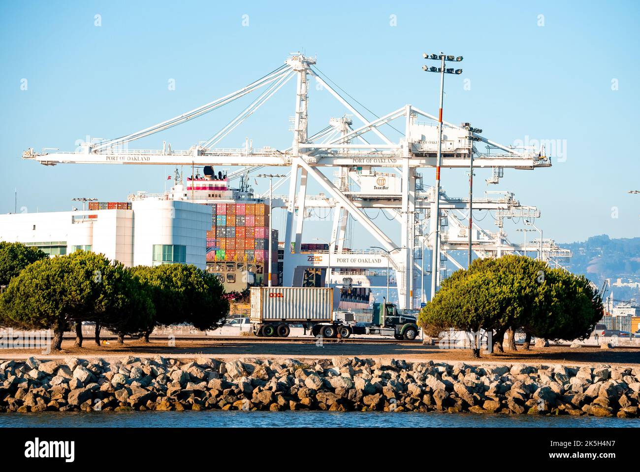 Loaded trucks moving by container cranes at Port of Oakland by bay during summer Stock Photo
