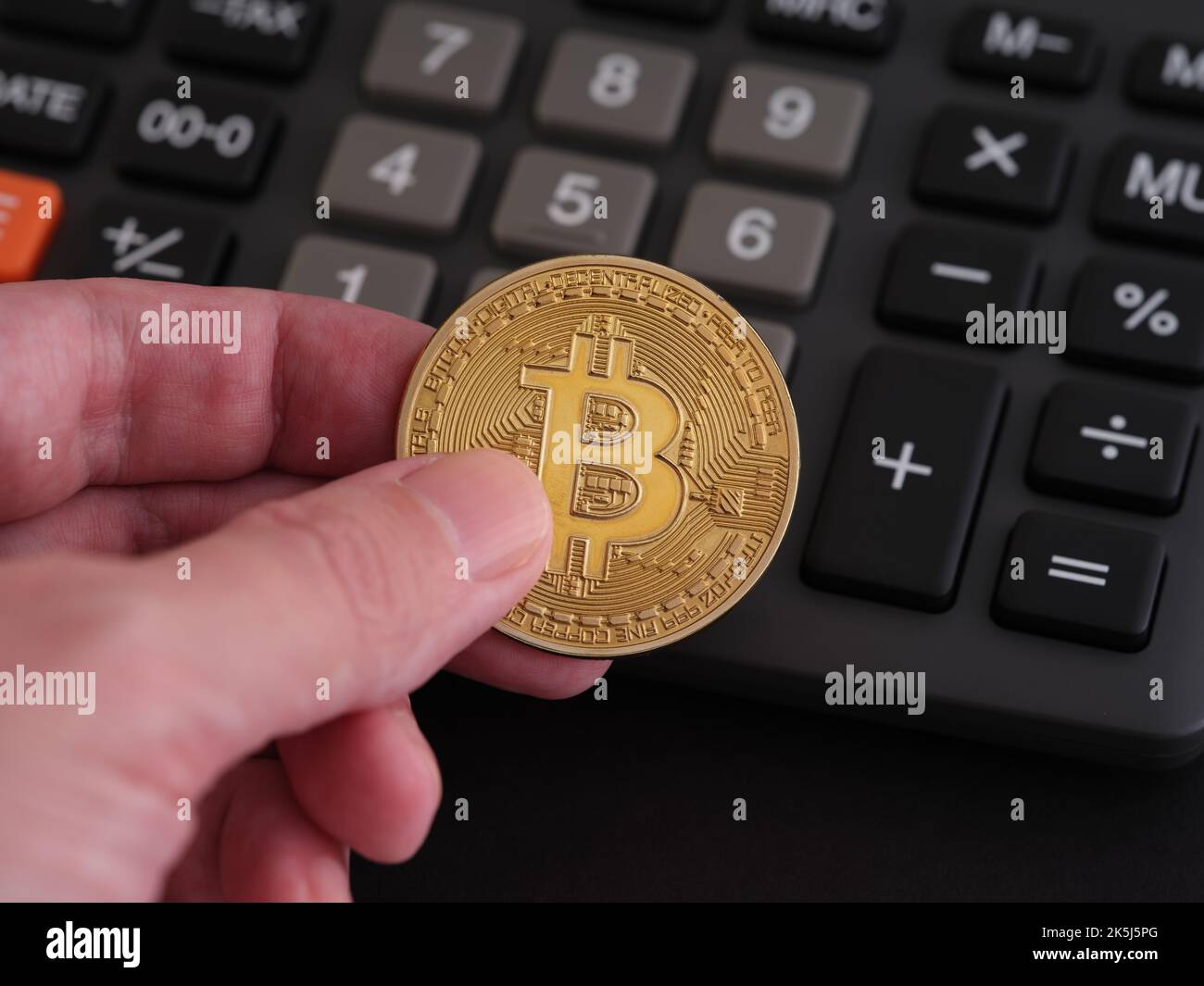 A man holding a golden bitcoin coin in his hand with a calculator in the background. Close up. Stock Photo