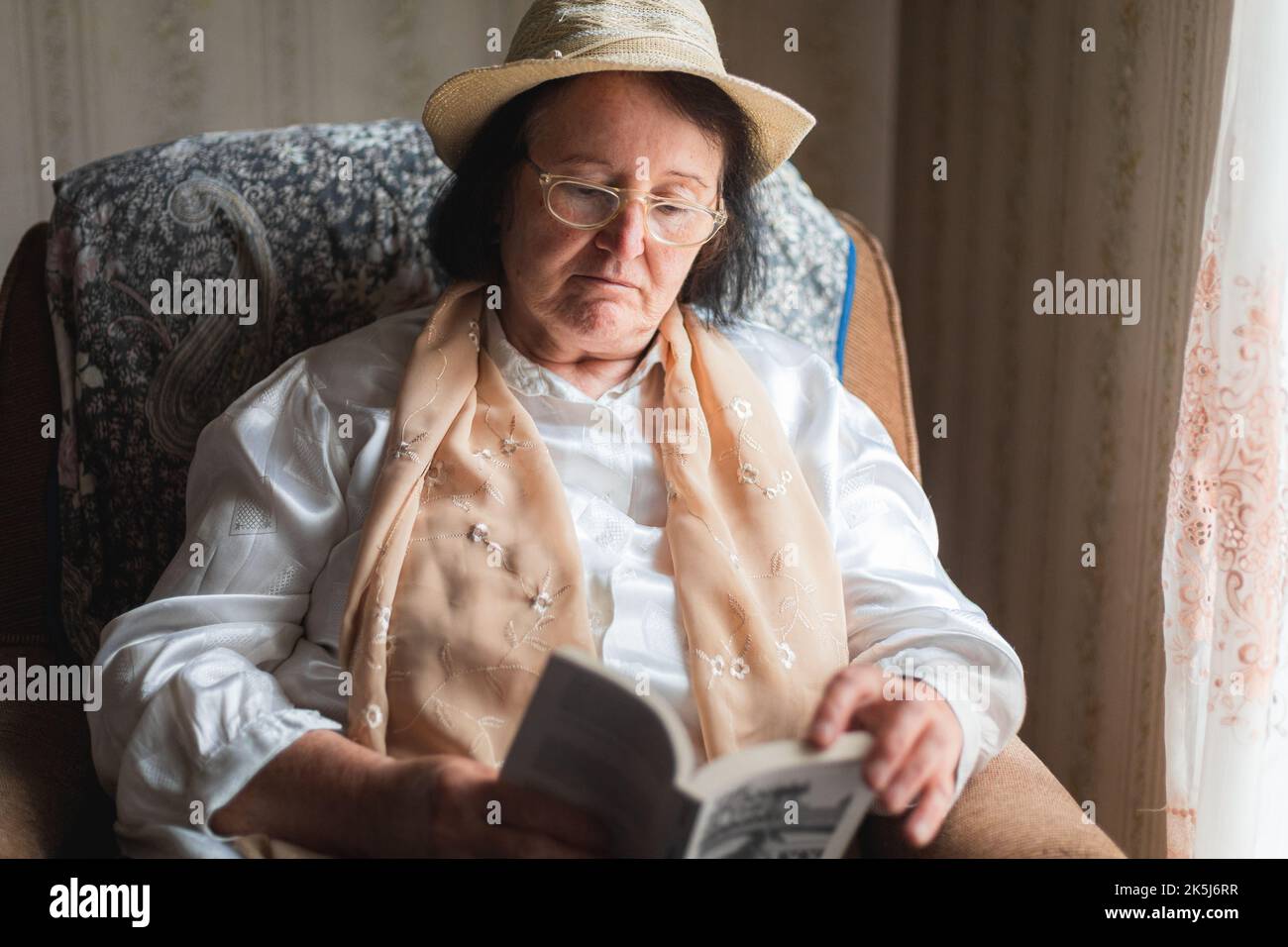 Elderly woman reading a book, sitting in the chair by the window Stock