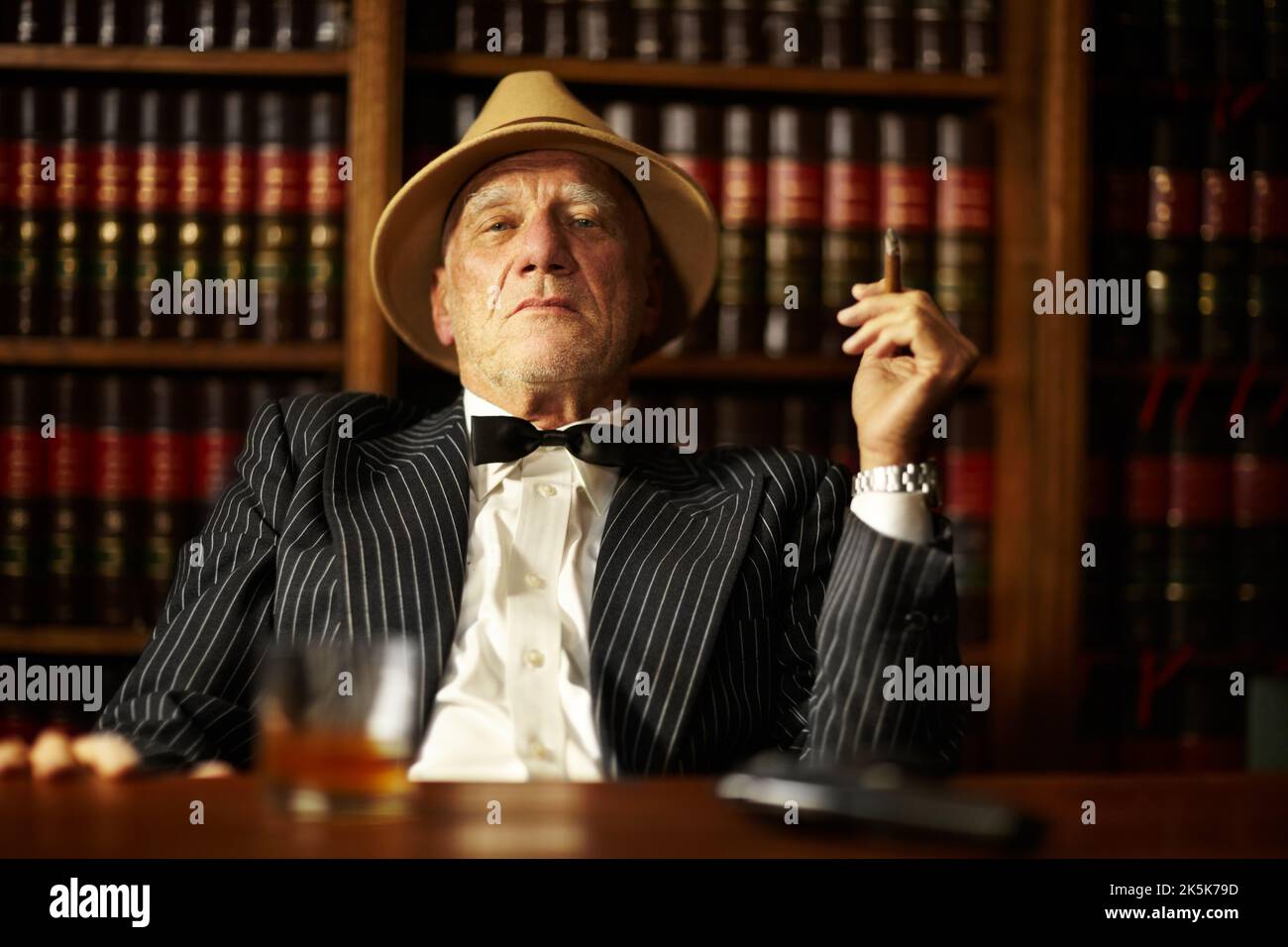 Crime is all about control. Aged mob boss smoking a cigar and looking serious from behind his desk. Stock Photo