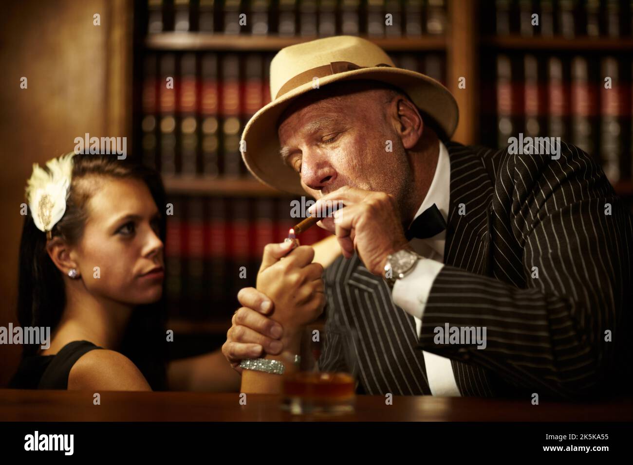 Hes in control of everyone around him. Aged mob boss wearing a hat and looking serious while a woman lights up a cigarette for him. Stock Photo