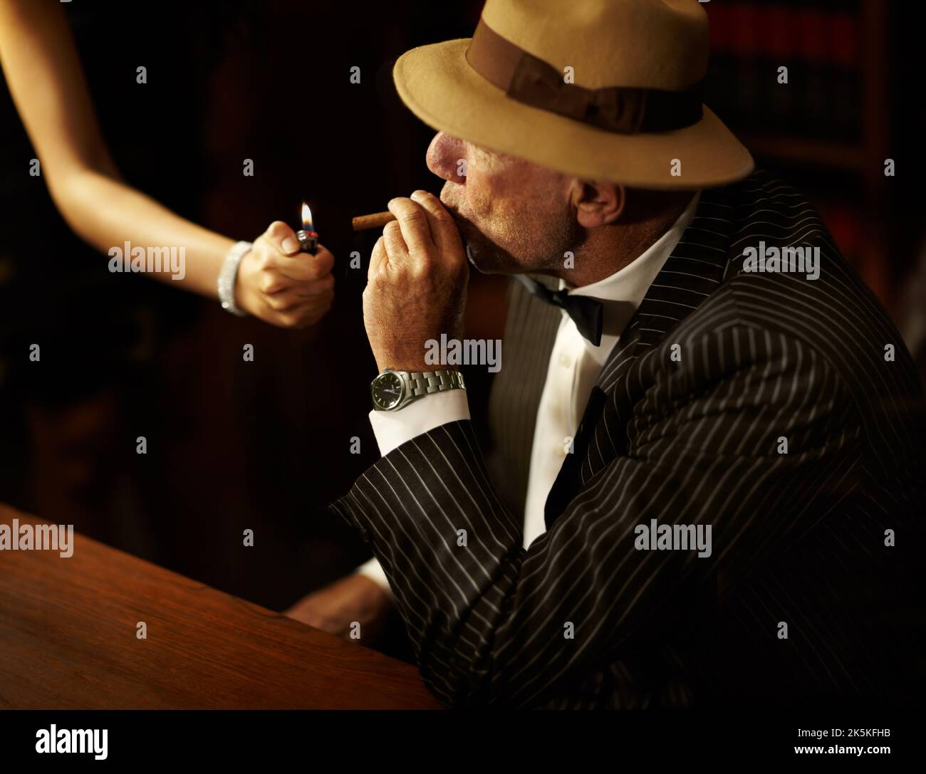 Hes got power and influence over others. Aged mob boss wearing a hat and looking serious while a woman lights up a cigarette for him. Stock Photo