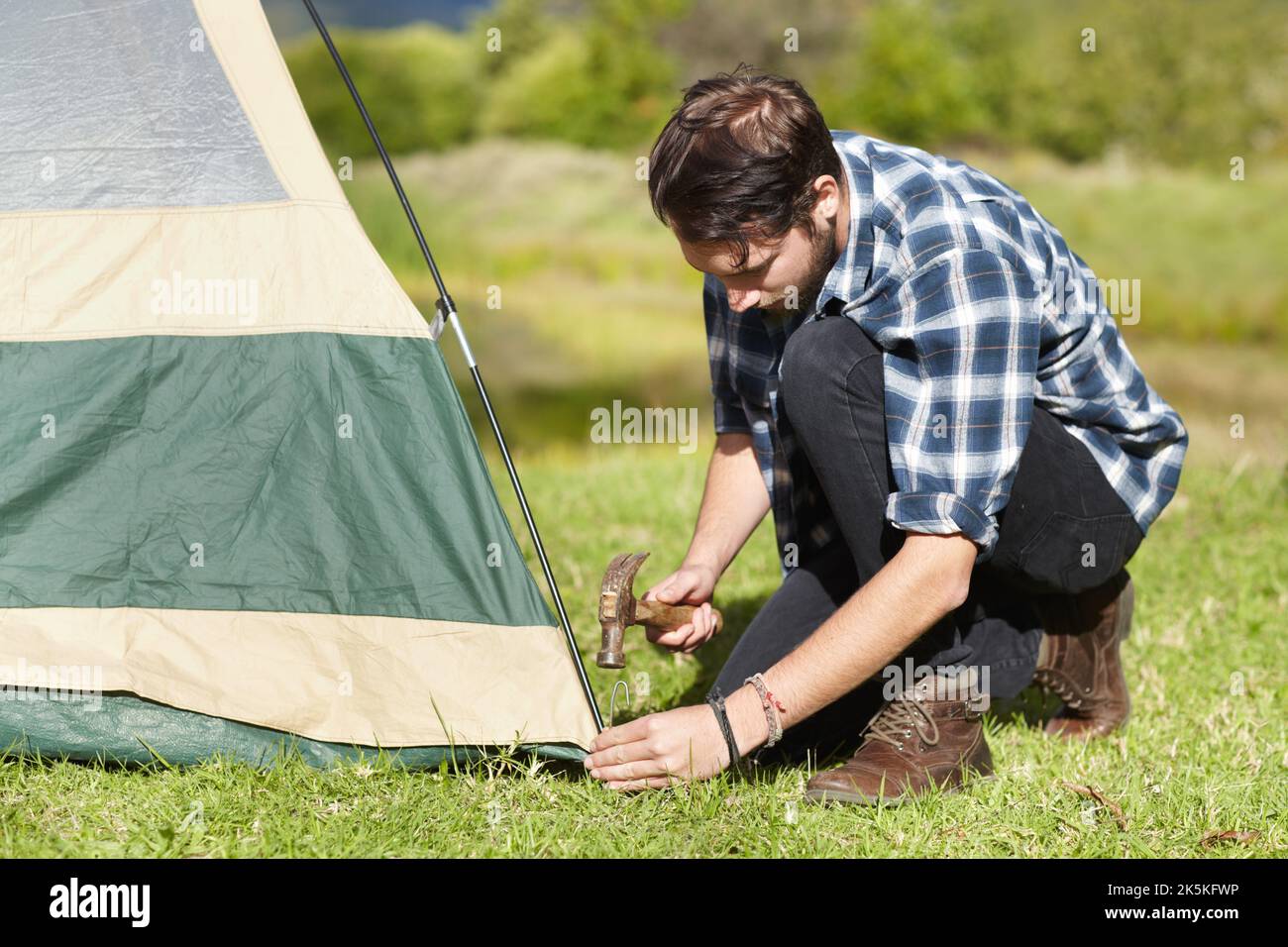 Better make sure its securely fastened. Handsome young man hammering a tent peg into the ground. Stock Photo