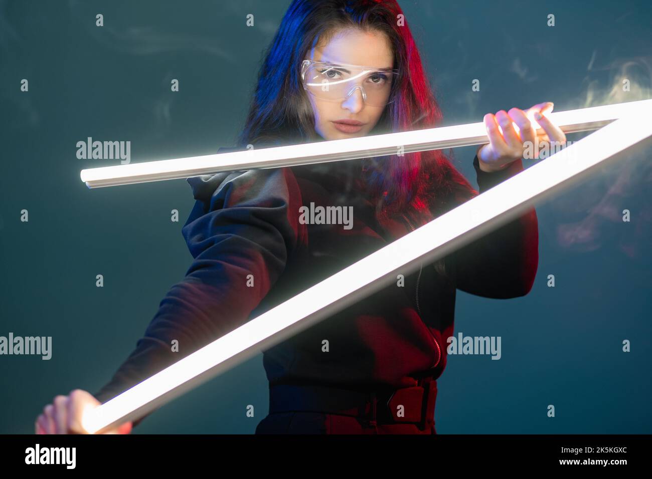 Cyberpunk hero. Futuristic portrait. Fantastic warrior. Red neon light brave female assassin in glasses ready for attack with white LED laser weapon i Stock Photo