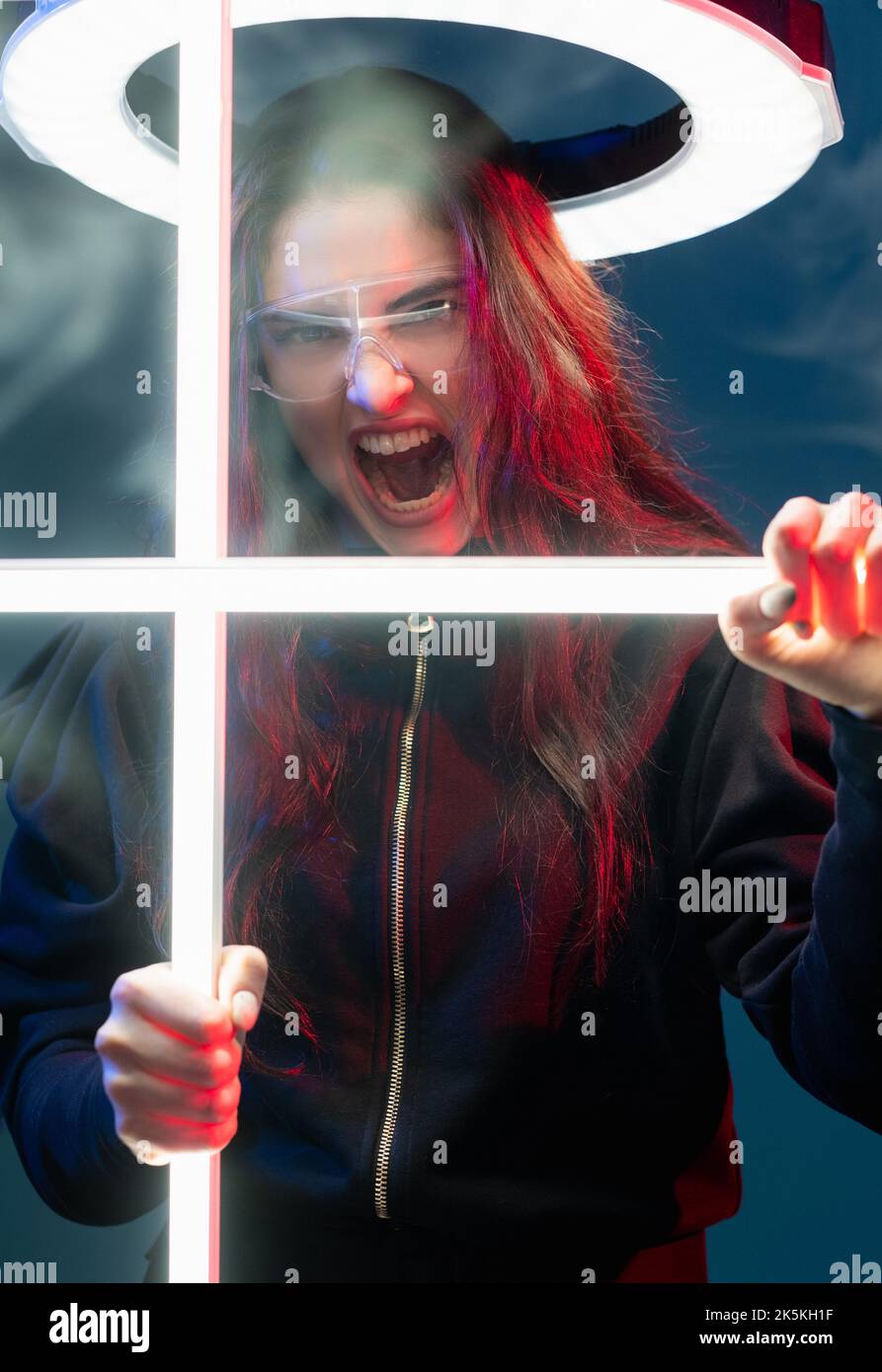 Mad fighter. Cyberpunk apocalypse. Futuristic attack. Frustrated angry red neon light cyborg woman in glasses yelling in white LED halo ring behind cr Stock Photo