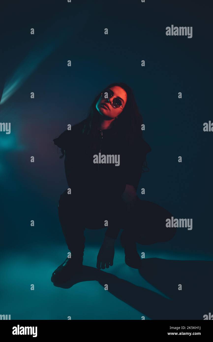 Urban girl. Night subculture. Underground style. Tough arrogant red neon light woman in black in sunglasses sitting on haunches on dark blue. Stock Photo
