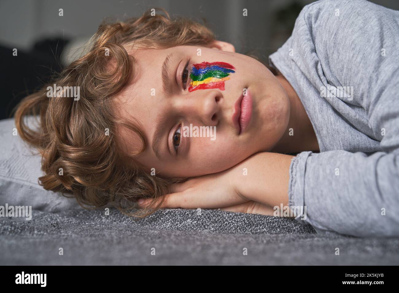 Unemotional boy with rainbow painted under eye lying on floor Stock Photo