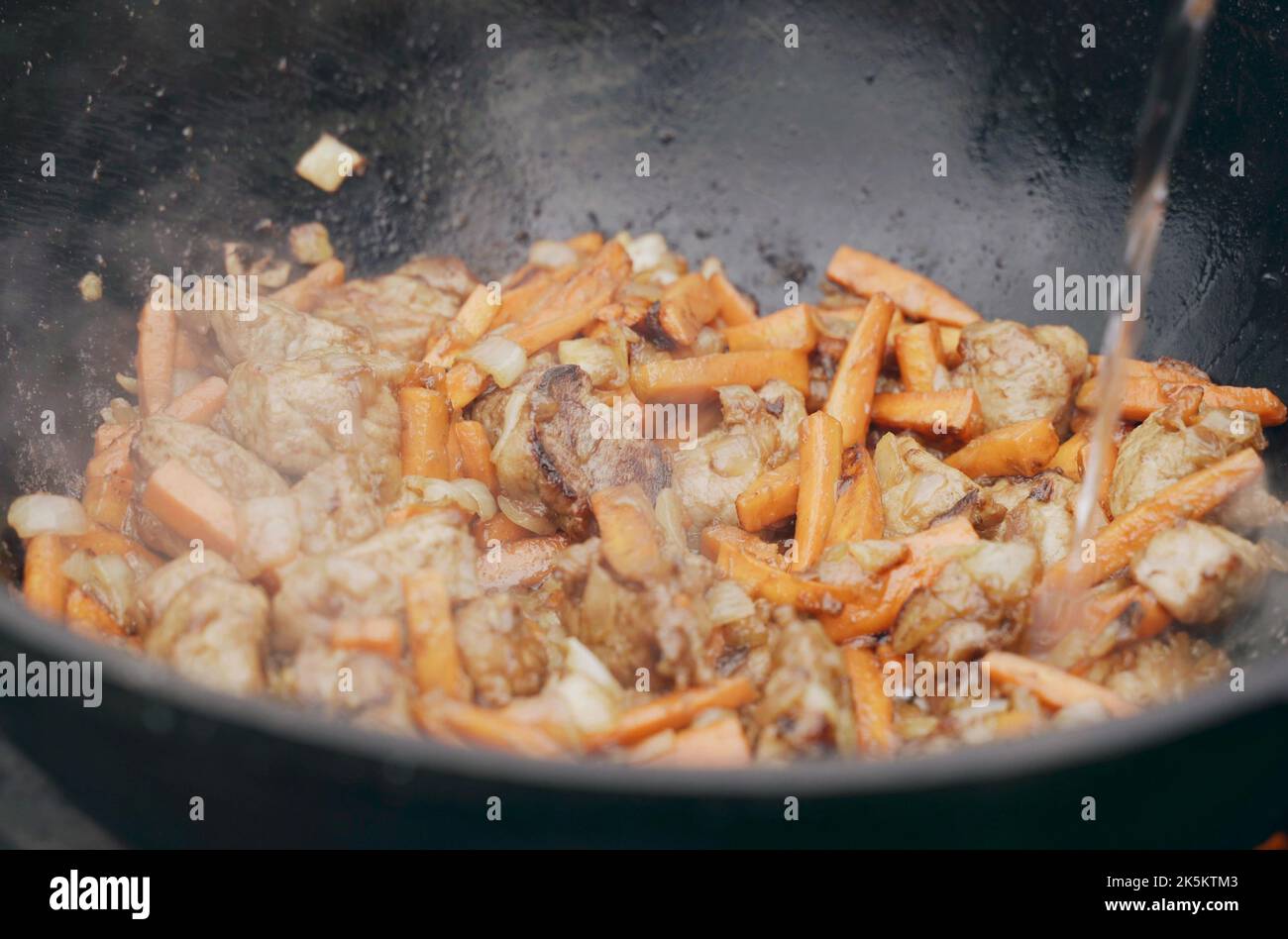 Water is poured into the cauldron where onions and meat and carrots are fried. Close-up. Stock Photo