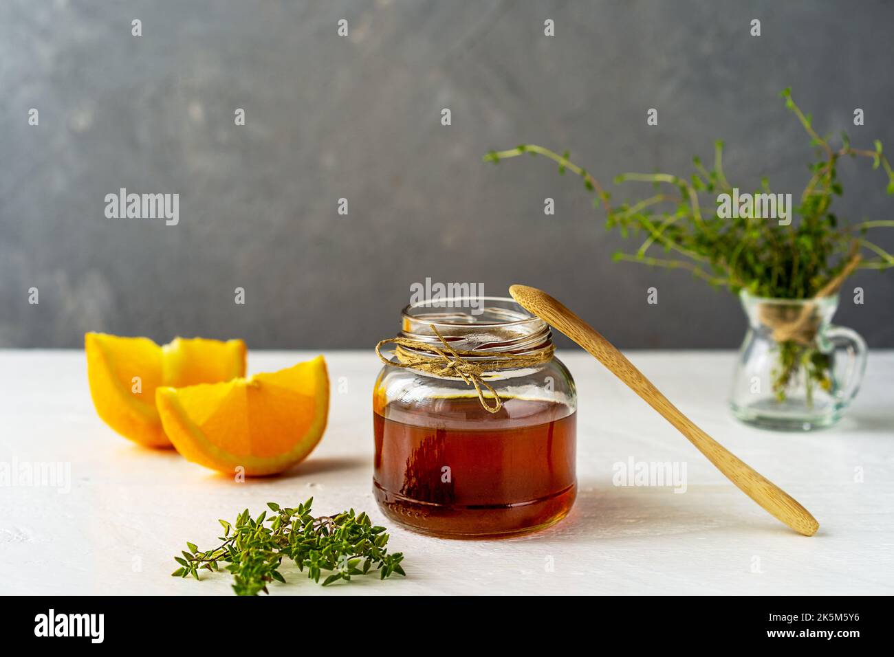 Thyme honey in small jar, wooden tea spoon, small thyme branch, orange slices on white table with grey background Stock Photo