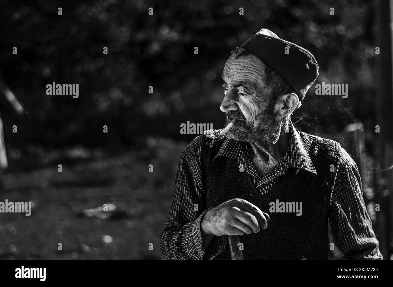 A grayscale portrait of an old man with a sajkaca hat looking aside, smoking in Valjevo, Serbia Stock Photo