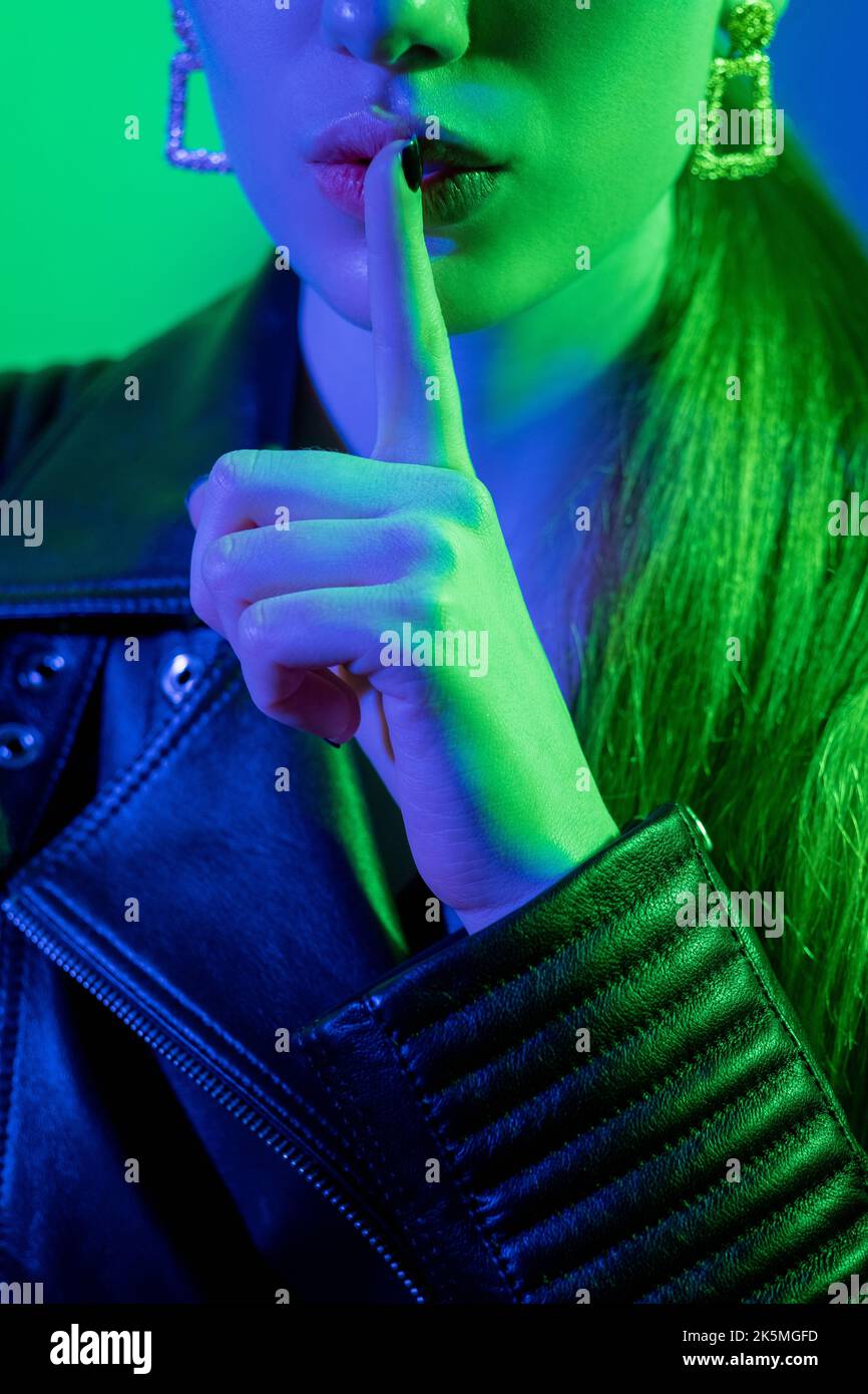 Hush gesture. Color light woman. Keep quiet. Futuristic secret. Mysterious girl in black leather jacket warning with shh finger on lips in green blue Stock Photo