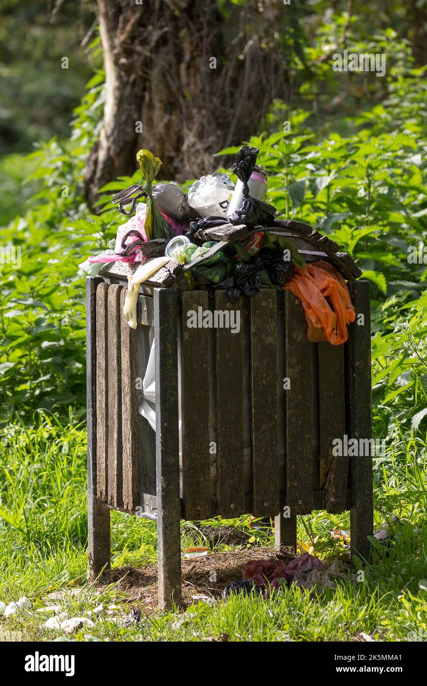 Rubbish bin full and overflowing along riverside walk. Full of food packaging drink cans and bottles dog poo bags and varoious other waste in portrait Stock Photo