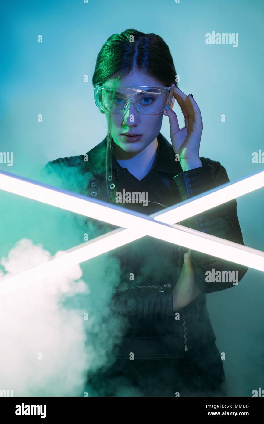Cyberpunk portrait. Color light people. Sci-fi future. Blue green neon light curious woman in protective goggles in white smoke cloud LED lamp glow. Stock Photo