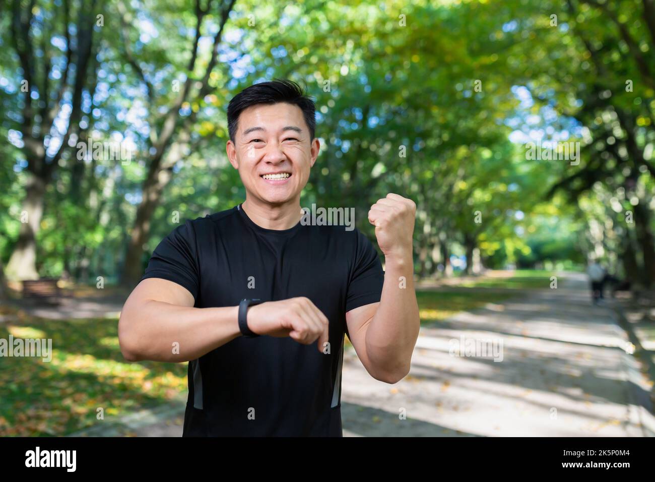 Happy and successful man looking at camera and smiling, asian sportsman satisfied with training result, using fitness bracelet smartwatch, holding hand up celebrating victory, success gesture. Stock Photo