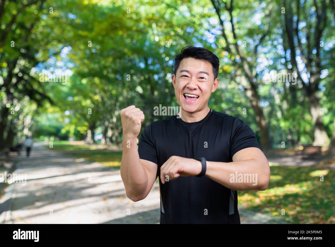 Happy and successful man looking at camera and smiling, asian sportsman satisfied with training result, using fitness bracelet smartwatch, holding hand up celebrating victory, success gesture. Stock Photo