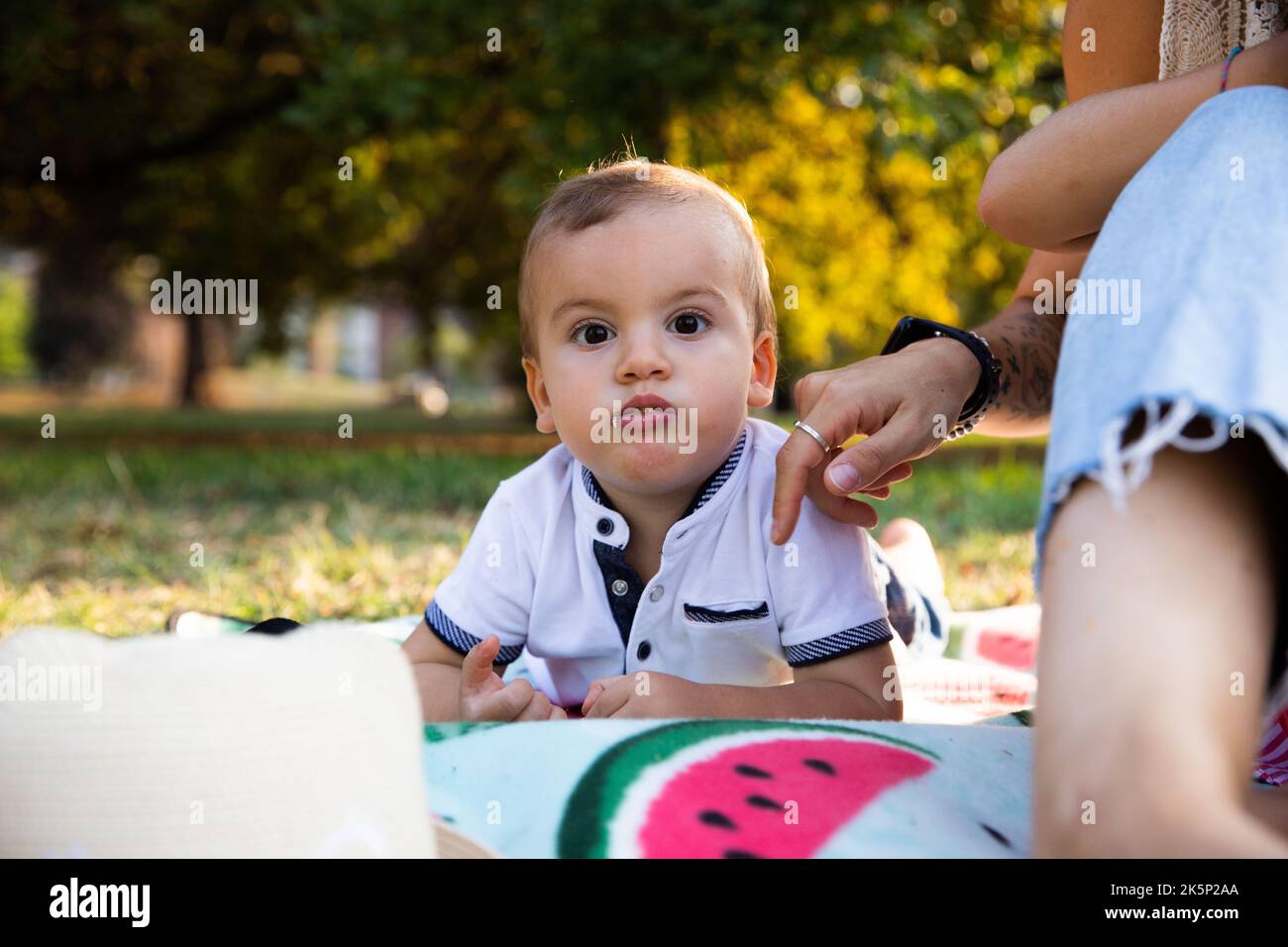 Toddler lying on picnic blanket with mouth full is watched over by mother sitting beside him. Stock Photo