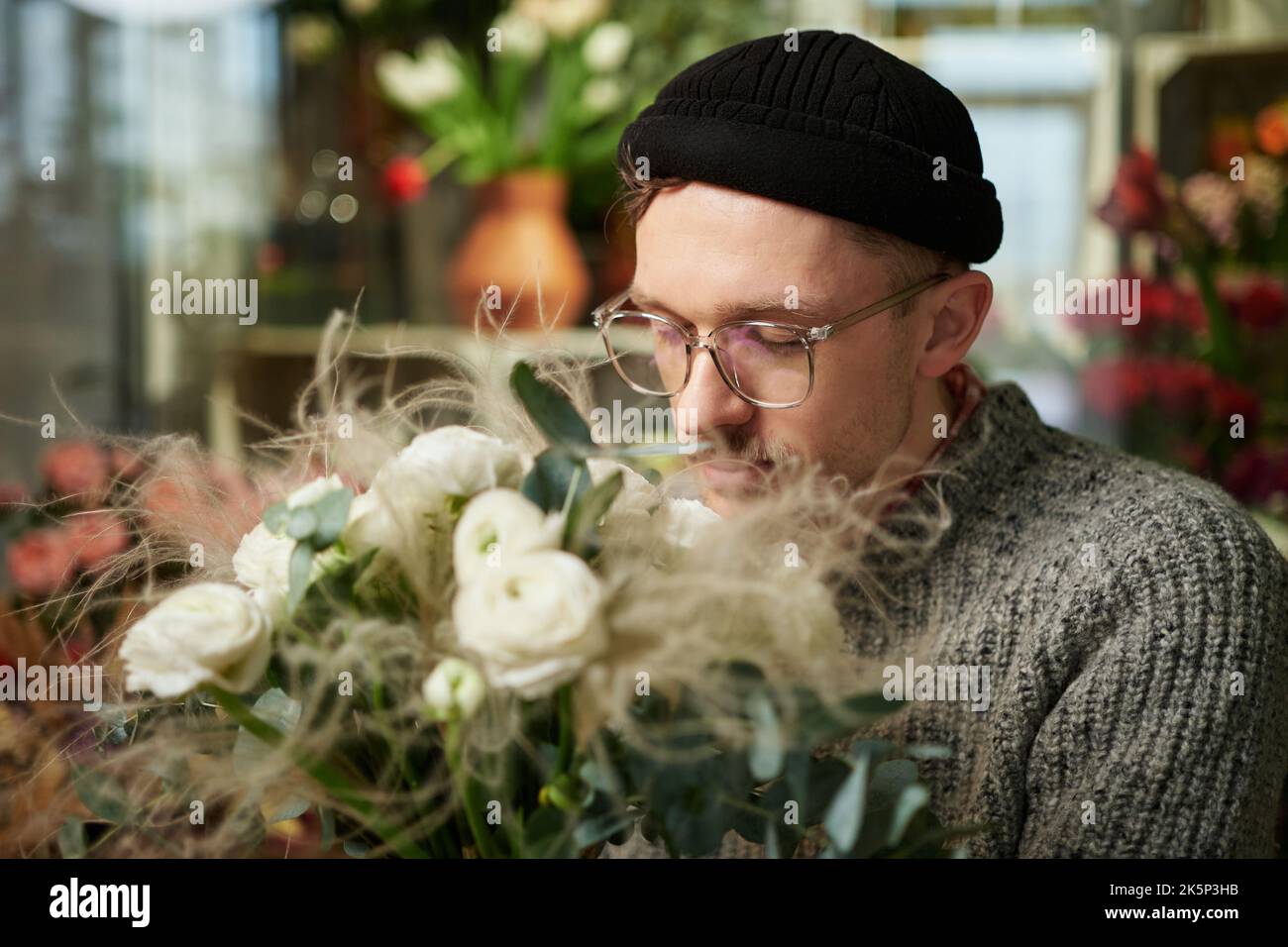 Florist male sniffing white roses bouquet in flower shop. Good looking male florist wearing sweater, beanie and eyeglasses with flower bouquet. Small business concept. High quality image Stock Photo