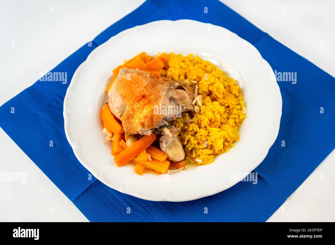 Baked chicken quarter, roasted carrot and stewed turmeric and saffron rice on white plate, blue towel on white table, closeup. Stock Photo