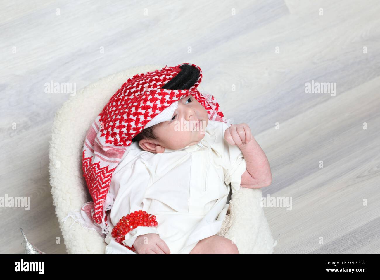 Saudi Muslim 1 month newborn boy in traditional arabian dressing white thobe red shemagh black agal sitting on little soft chair cultural tiny shoes Stock Photo