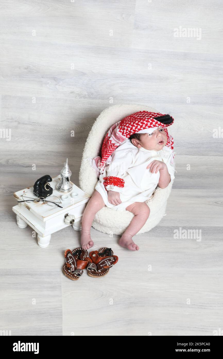Saudi Muslim 1 month newborn boy in traditional arabian dressing white thobe red shemagh black agal sitting on little soft chair cultural tiny shoes Stock Photo