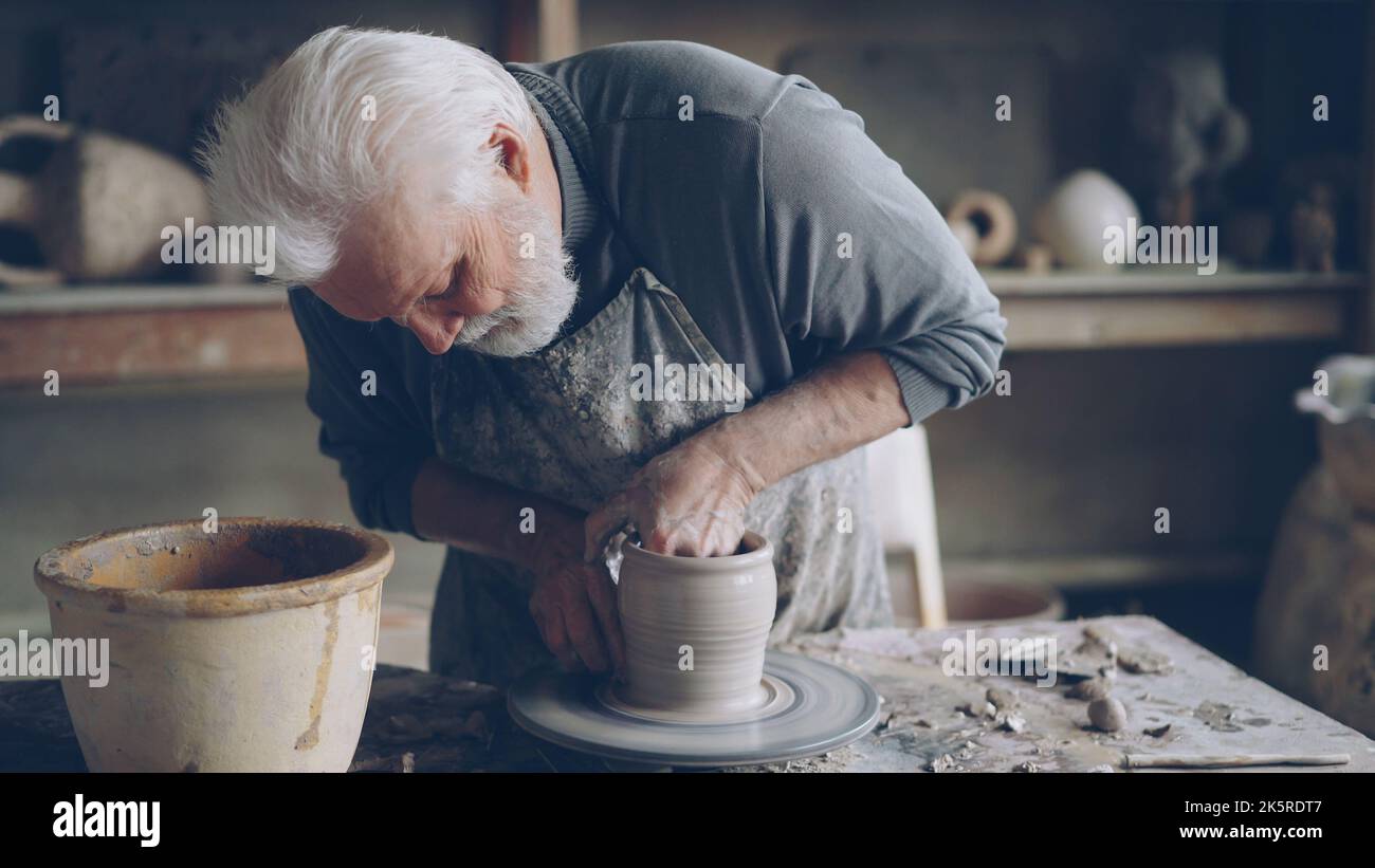 Experienced ceramist grey-haired bearded man is smoothing molded ceramic pot with wet sponge. Spinning throwing wheel, muddy work table and handmade clayware are visible. Stock Photo