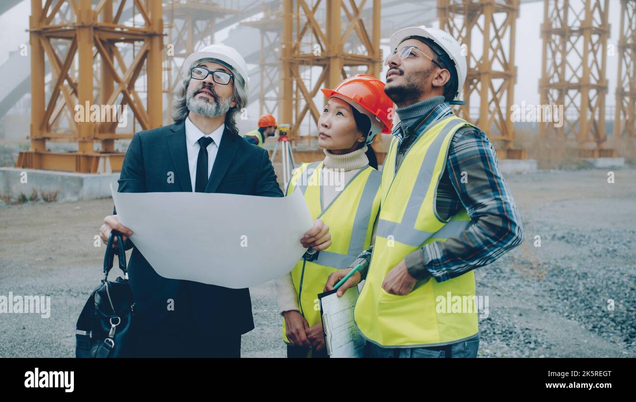 Architect mature man in suit is discussing project with builders wearing uniform showing blueprint standing at construction site. People and job concept. Stock Photo