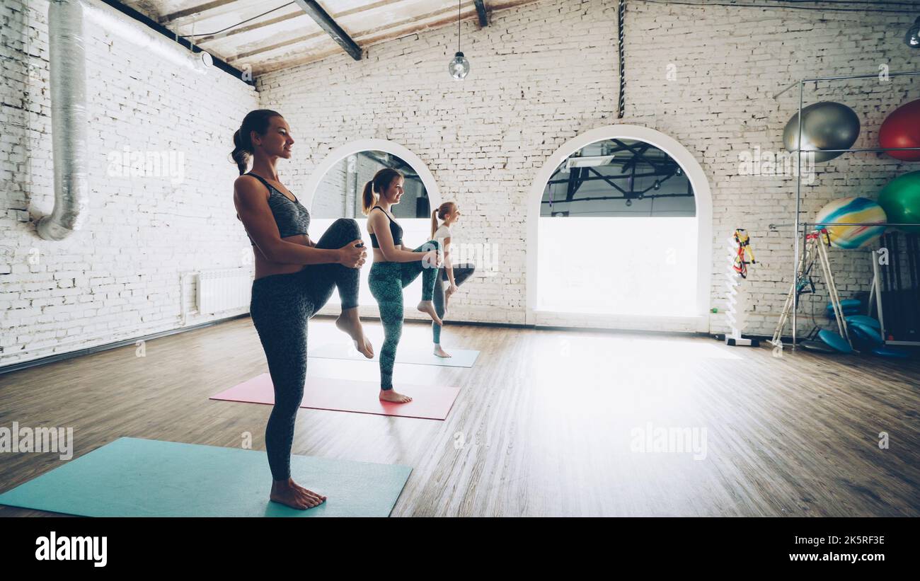Dolly shot of pretty slim young women doing balancing yoga exercises in light loft style gym. Students are concentrated on practice. Sports equipment, fitness balls and mats are visible. Stock Photo
