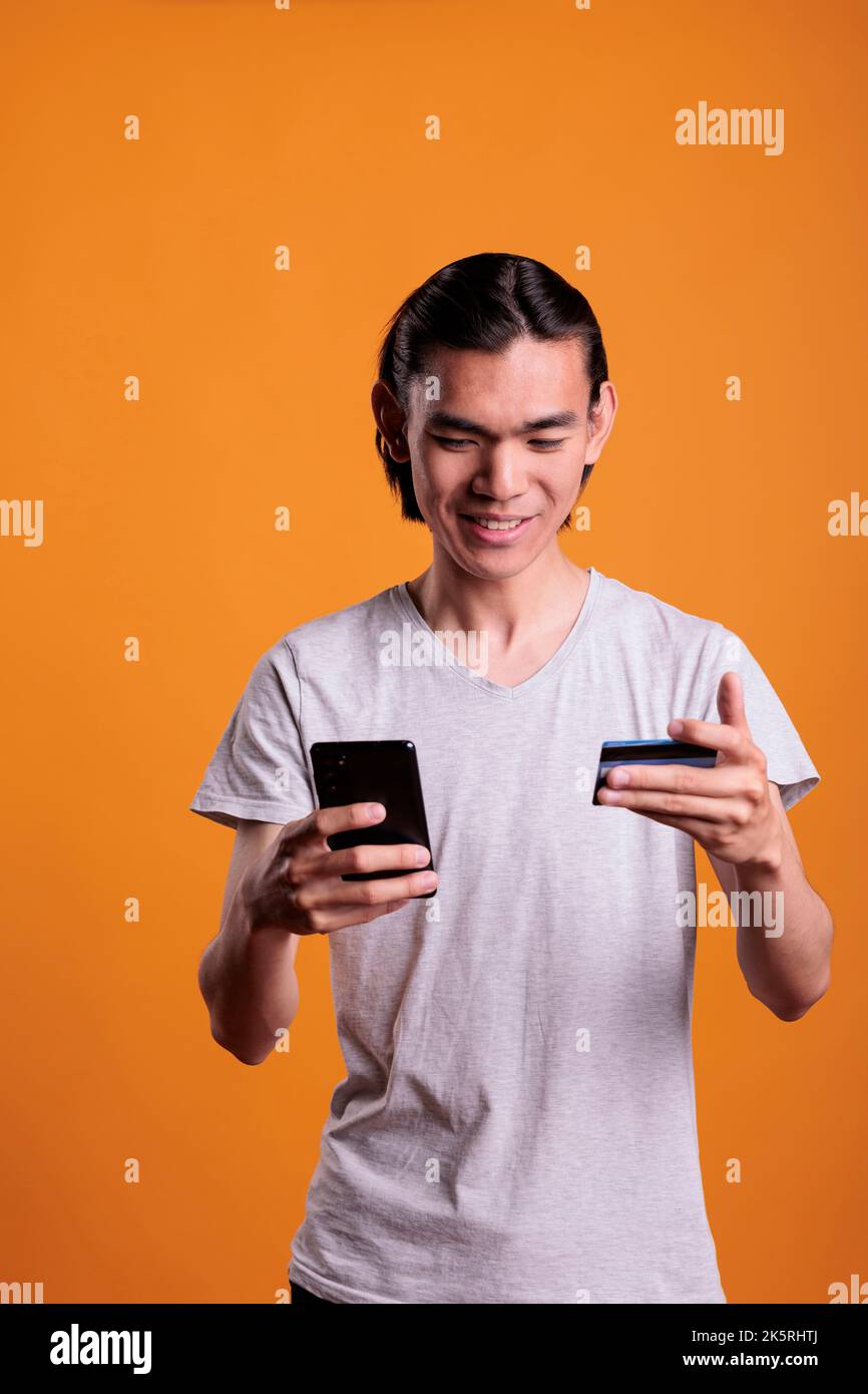 Smiling young man making order in internet shop, holding smartphone and credit card. Asian cheerful teenager buying products, searching goods in online store, e commerce concept Stock Photo