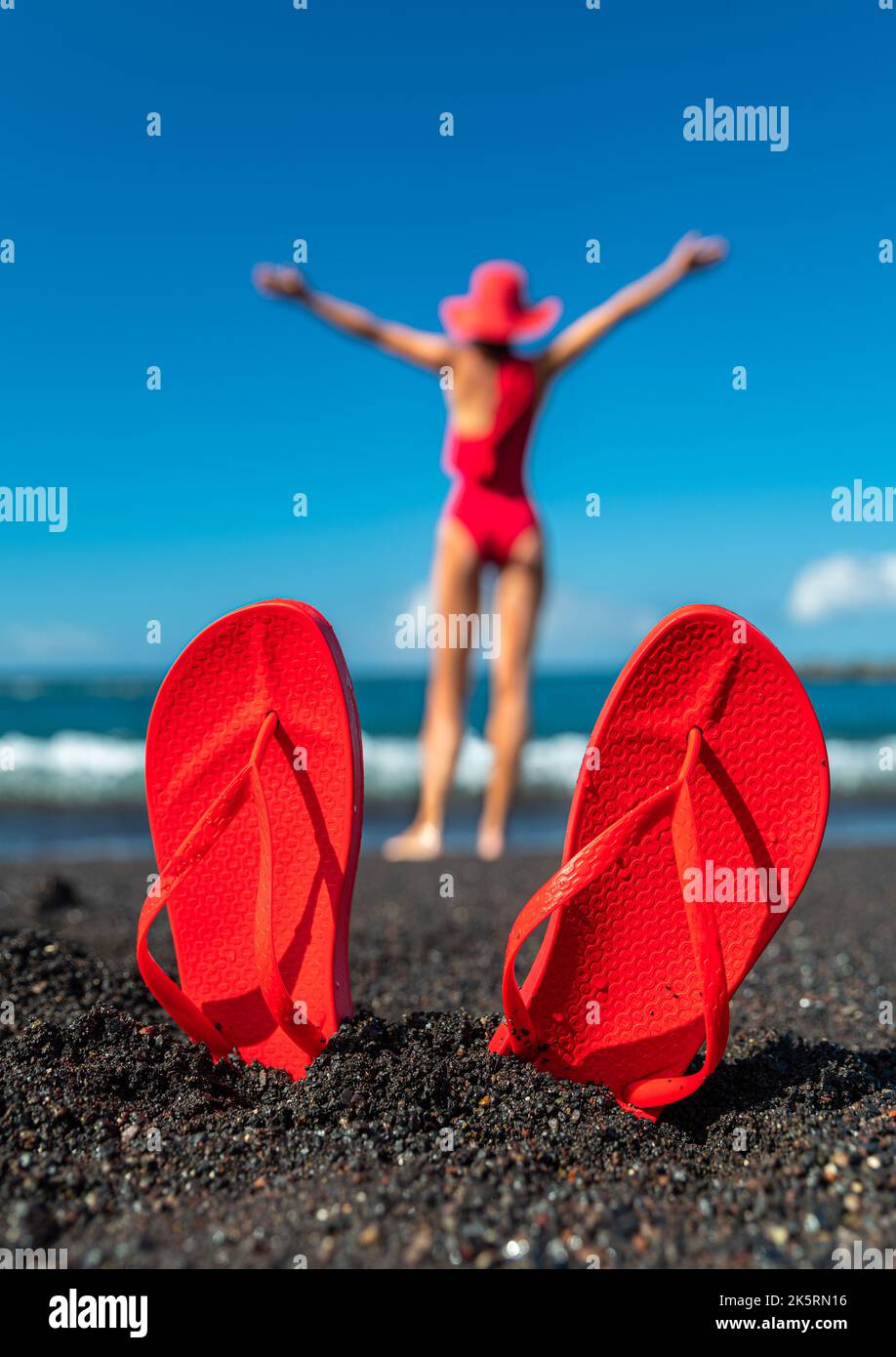 Red flip flops in the black sand and silhouette of woman in red swimsuit standing on the ocean beach. Summer concept. Stock Photo