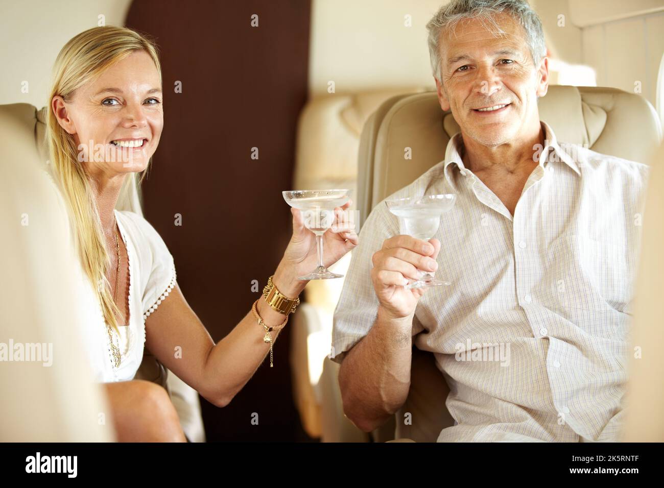 Cheers. Smiling couple seated in a private jet and toasting each other - portrait. Stock Photo