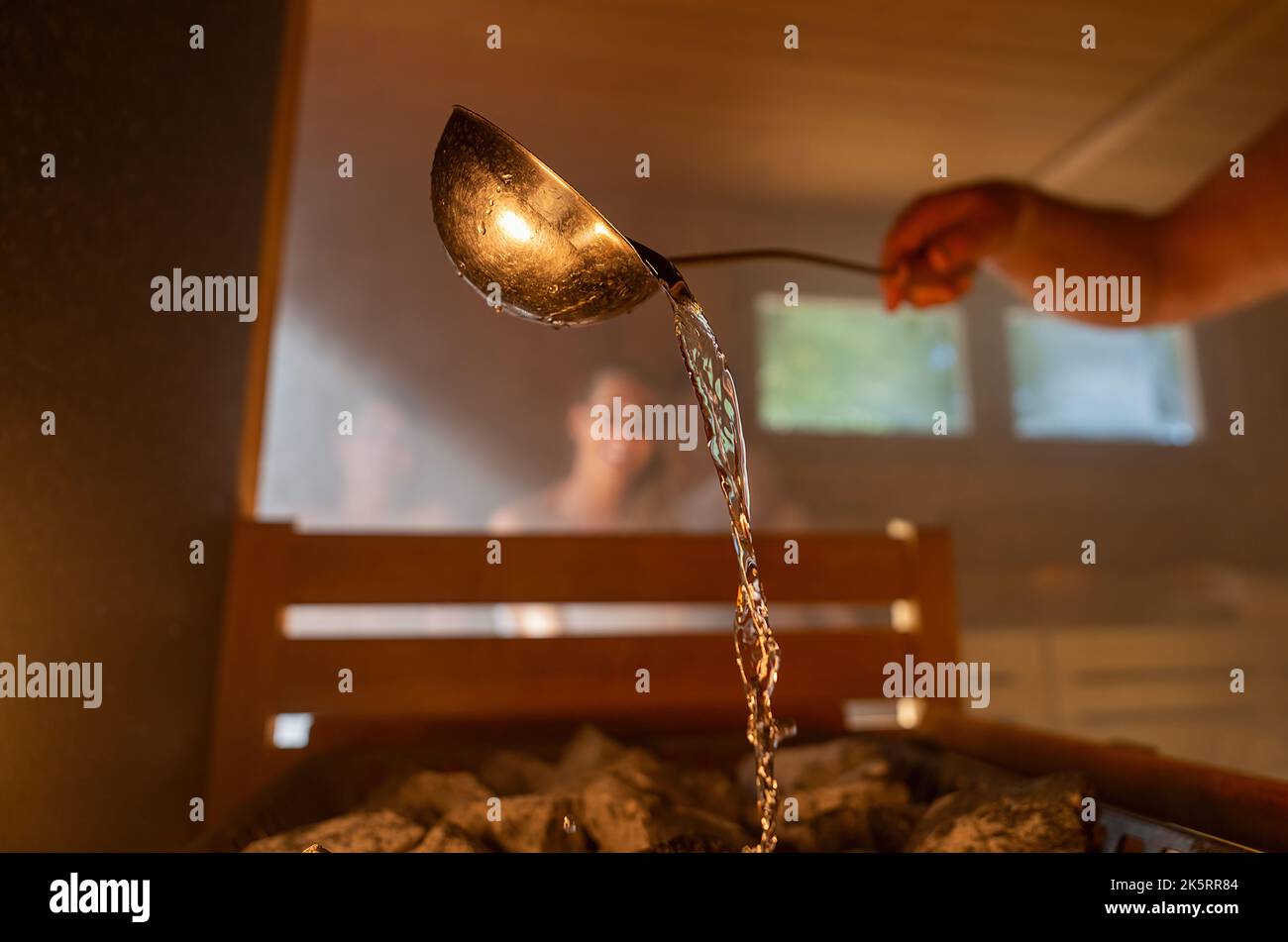 Man pouring water onto hot stone with metal spo in sauna room with a group of people. Steam an water on the stones, spa and wellness concept, relax in Stock Photo