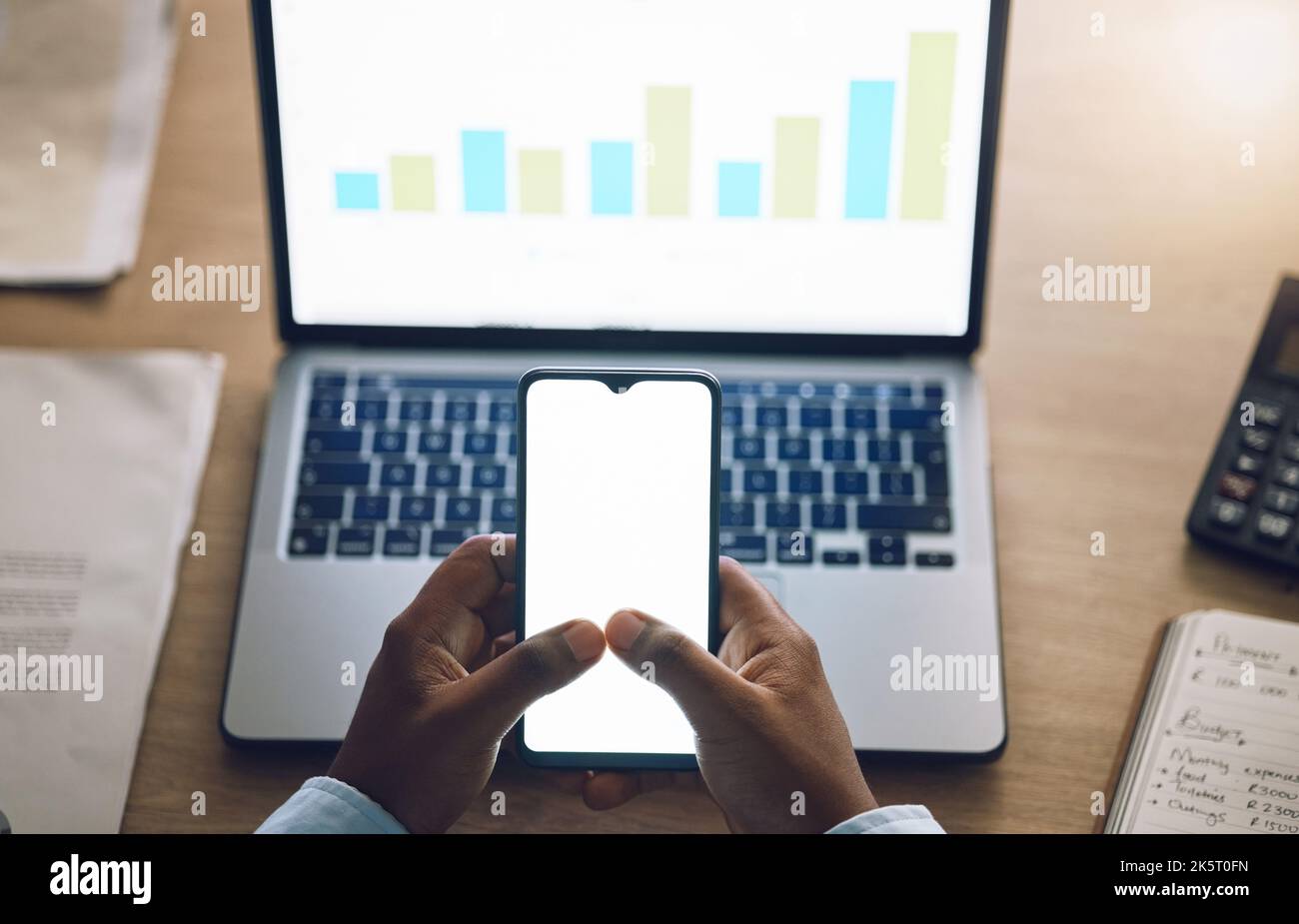 Above closeup hands of african man using a phone while working on his laptop. African american business man looking at graphs while working late at Stock Photo