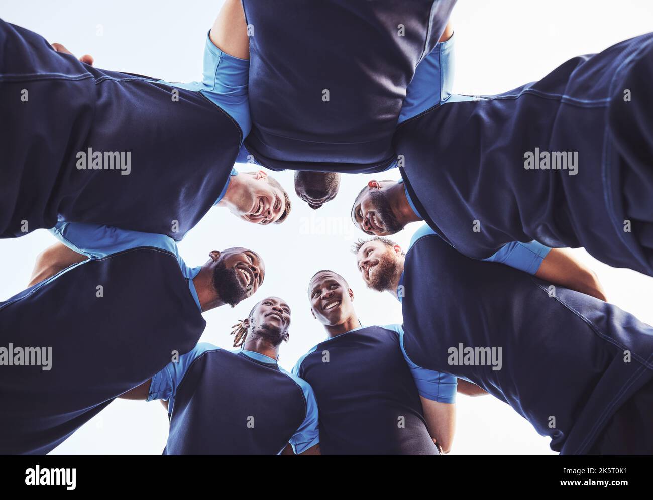 Below diverse group of rugby players standing in a huddle together outside on a field. Young male athletes looking serious and focused while huddled Stock Photo