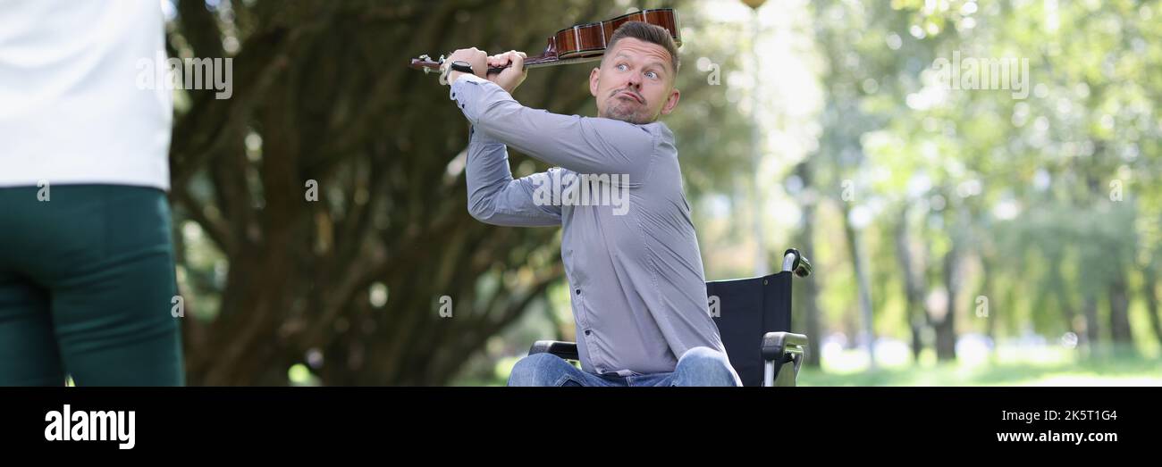 Man in wheelchair swings guitar at woman in park Stock Photo