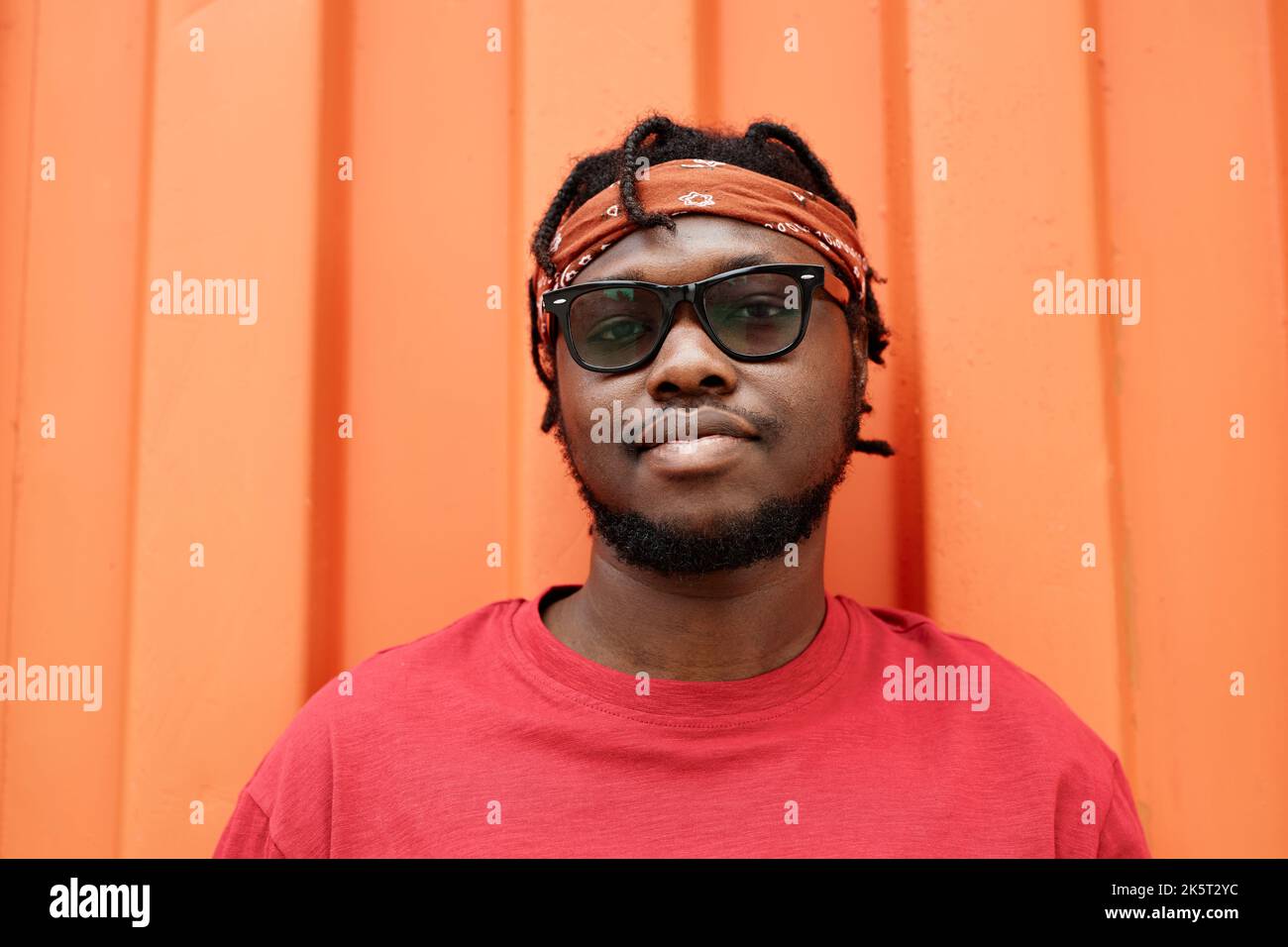 Front view portrait of young African American man looking at camera against red color wall in urban setting Stock Photo