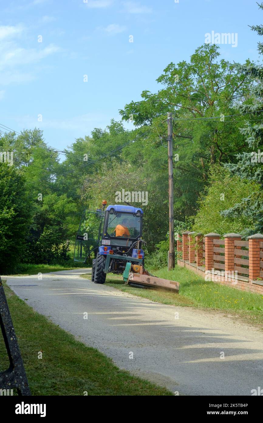 small municipal tractor owned by the local authorities being used to cut grass verges in rural lane of small hamlet zala county hungary Stock Photo