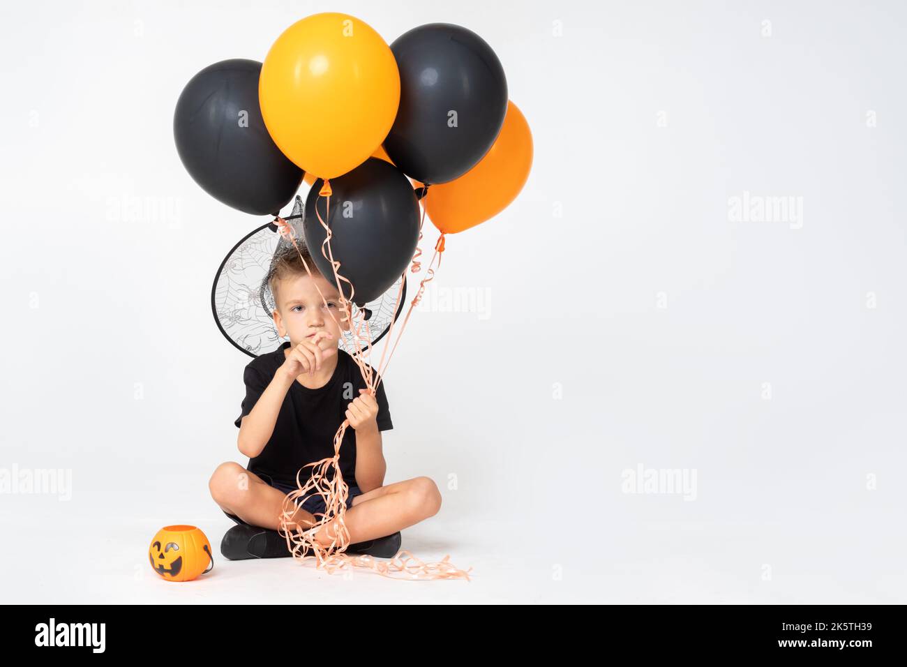 A cheerful happy boy in a wizard's hat holding a pumpkin-shaped bucket and black and orange balloons, sitting on a white studio background. Ready for Stock Photo