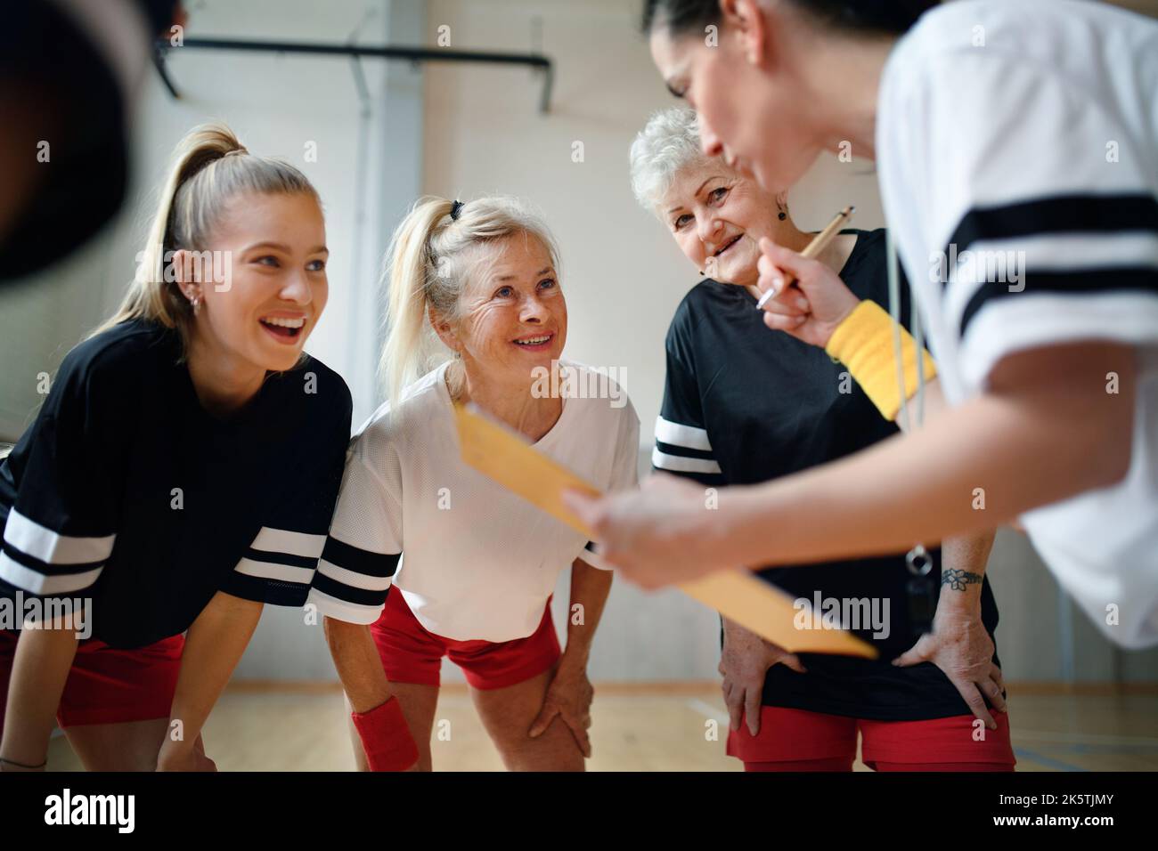 Female sport coach with clipboard discussing tactics with young and old women team training for match in gym. Stock Photo