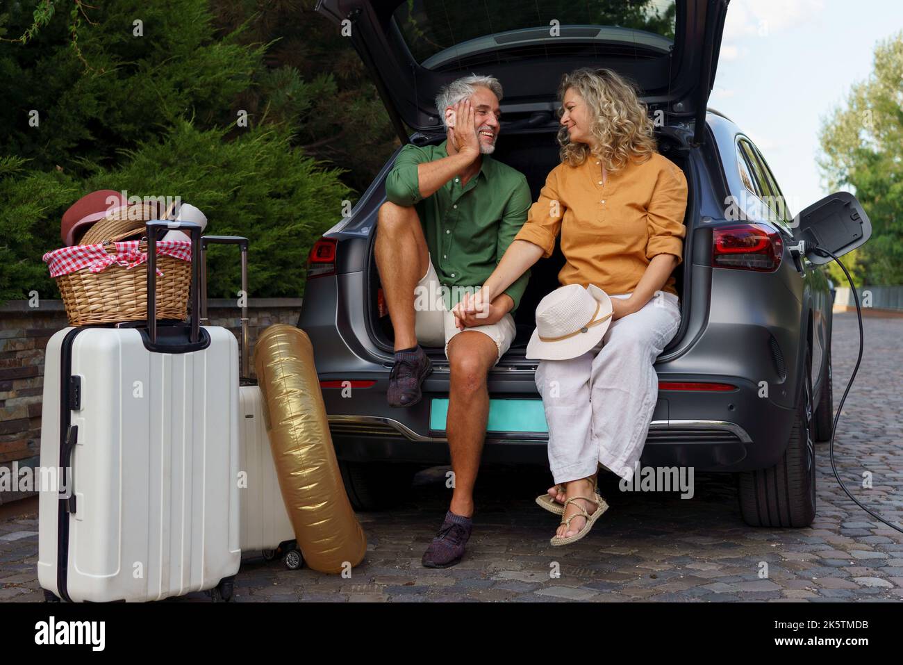 Middle-aged couple sitting in trunk while waiting for charging car before travelling on summer holiday. Stock Photo