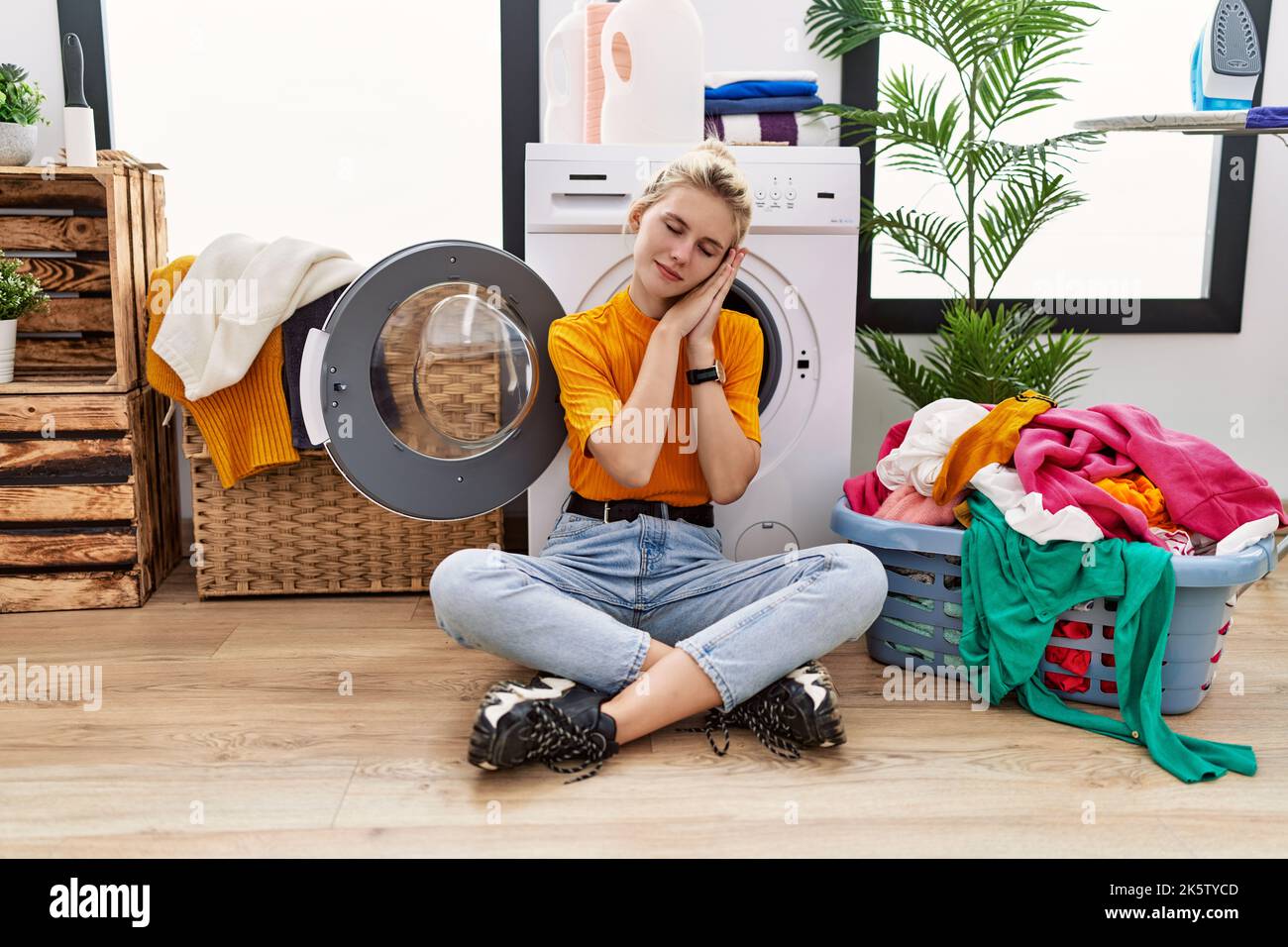 Young blonde woman doing laundry sitting by washing machine sleeping tired dreaming and posing with hands together while smiling with closed eyes. Stock Photo