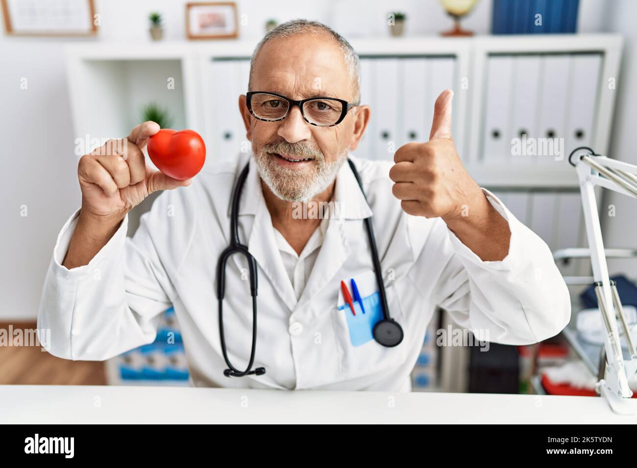 Mature doctor man holding red heart at the clinic smiling happy and ...