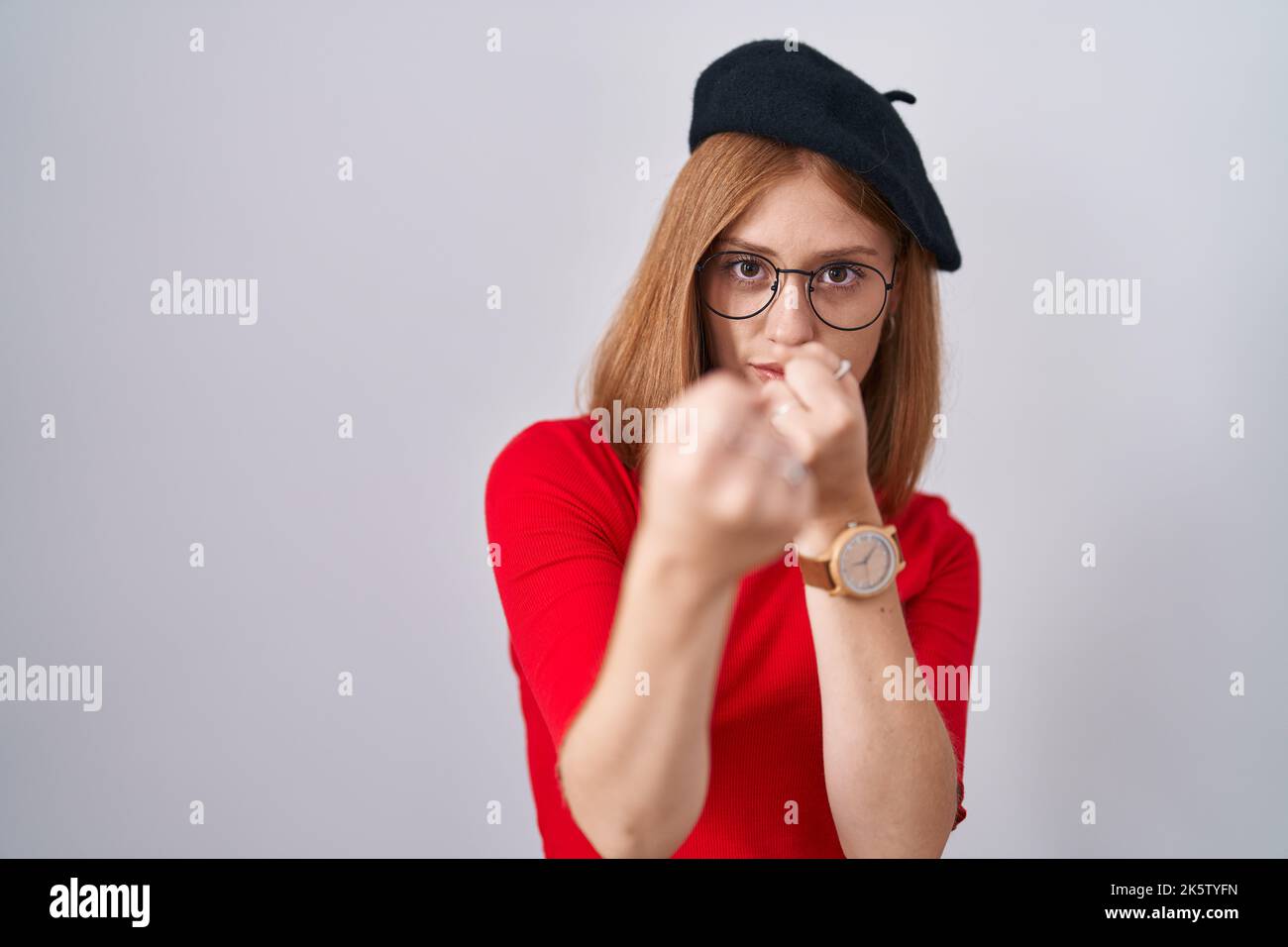 Young redhead woman standing wearing glasses and beret ready to fight with fist defense gesture, angry and upset face, afraid of problem Stock Photo