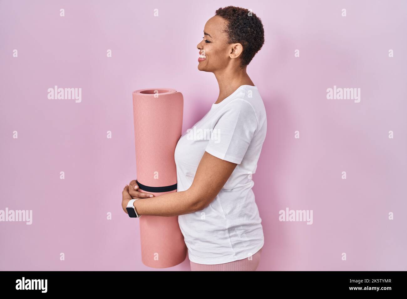 Beautiful african american woman holding yoga mat looking to side, relax profile pose with natural face and confident smile. Stock Photo