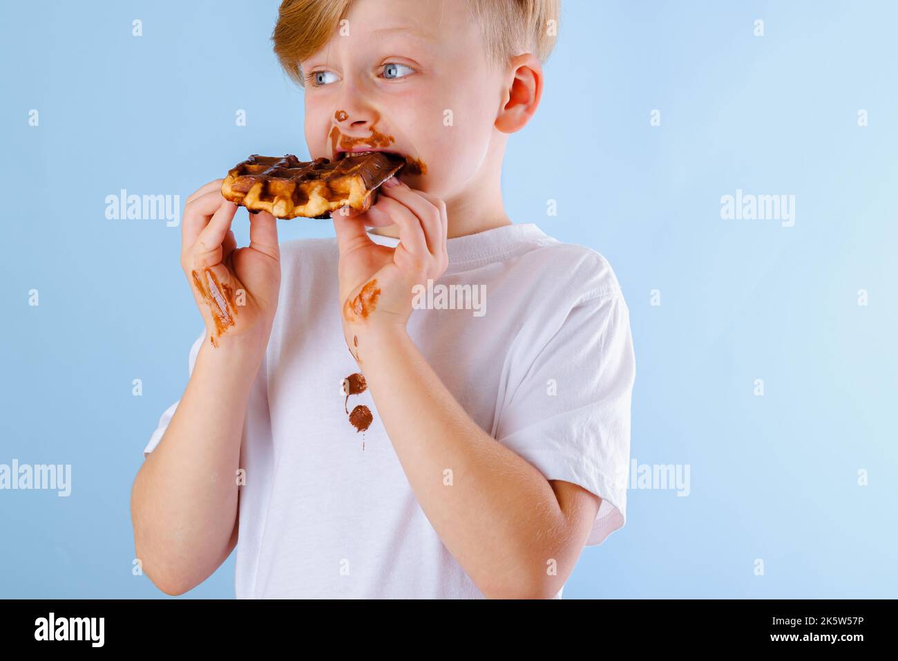 The child is eating Belgian waffle with chocolate sauce on a blue background. Dirty chocolate stains on clothes and hands Stock Photo