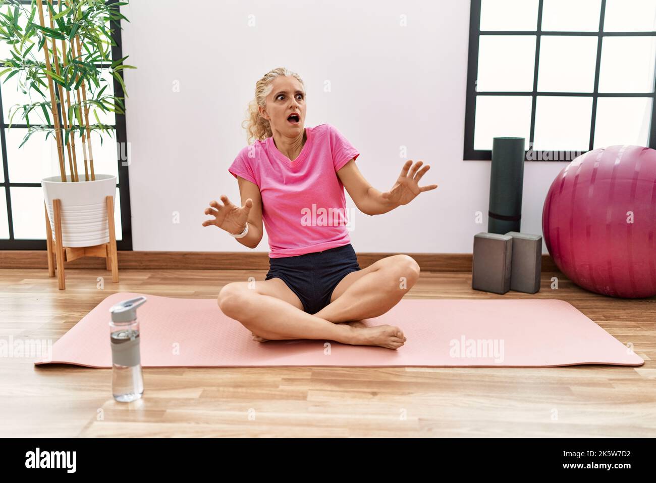 Middle age blonde woman sitting on yoga mat afraid and terrified with ...